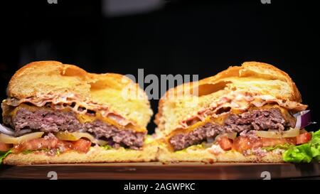 Cut craft burger is cooking on black background. Consist: sauce, lettuce, tomato, red onion, pickle, cheese, bacon, air bun and marbled meat beef. Stock Photo