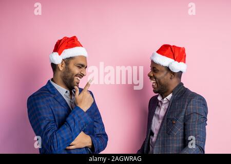 Joyful afro american entrepreneur and indian businessman smiling and laughing, standing over pink background, wearing Santa hats. Best friends having Stock Photo