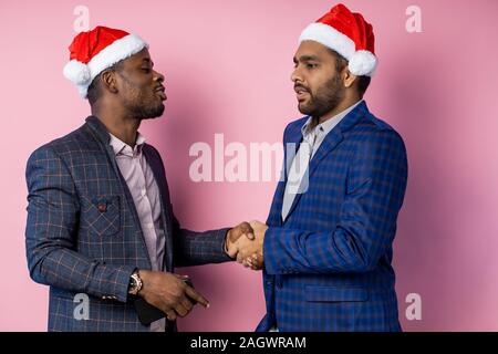 Portrait of two multicultural businessmen partners wearing formal clothing, Santa hats, shaking hands after making good deal, smiling, congratulating Stock Photo