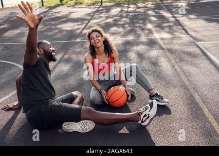 Outdoors Activity. African couple sitting with ball on basketball court man raising hand up laughing cheerful Stock Photo