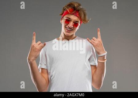 European blond guy with a bandana in glasses and a white T-shirt shows a rocker sign on a gray background Stock Photo