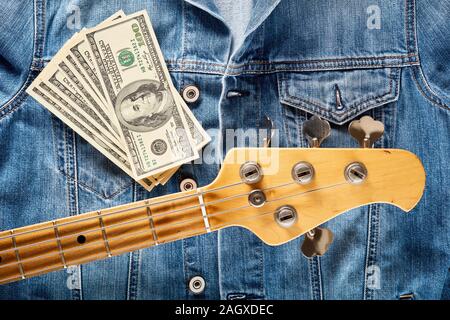 Bass guitar headstock and one hundred dollar bills on denim jeans jacket. Concept of  making or earning money in music industry. Stock Photo