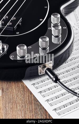 Black bass guitar and knobs with jack cable on sheet music and wooden table. Concept of music recording or composing. Close up view. Stock Photo