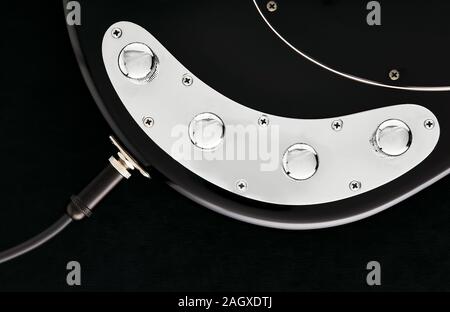 Cable jack connected to black electric bass guitar input with volume and tone control knobs. Close up detail on black background. Stock Photo