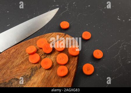 Carrot sliced to small circles on wooden chopping board, chef knife next black marble like desk. Stock Photo