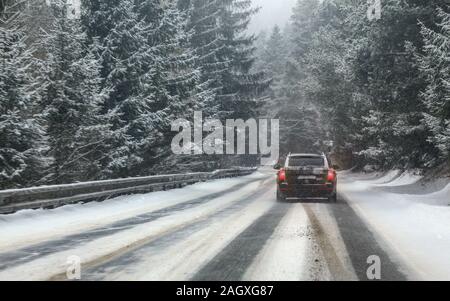 Black car drives on snow covered forest road during snowstorm, trees on both sides, view from car behind. Dangerous driving conditions. Stock Photo