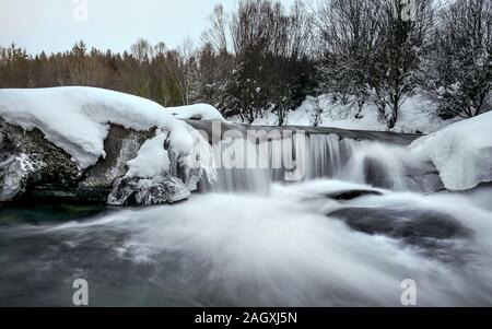 Small waterfall on river in winter, long exposure makes water milky smooth, snow and ice around, morning sun setting up distant sky. Stock Photo