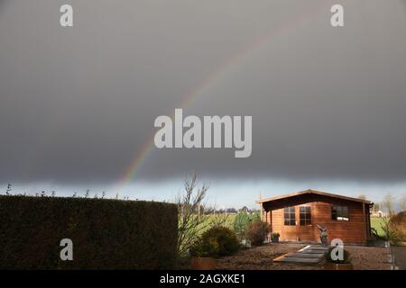 A rainbow over a dark grey sky in the garden. A wooden house at the background Stock Photo