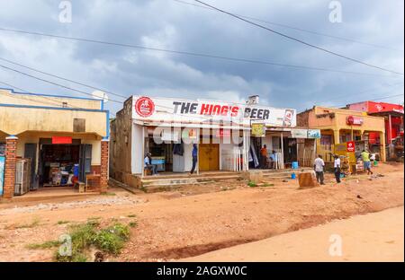 Typical low-rise roadside village shops advertising The King's Beer and buildings with local people in the Western Region of Uganda, on a cloudy day Stock Photo