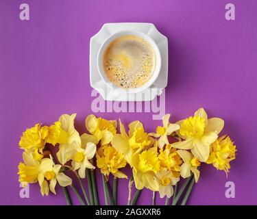 minimalistic flat lay with cup of coffee and yellow narcissus flowers on vibrant purple background Stock Photo