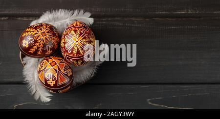 Pysanky on a small plate, Ukrainian Easter eggs decorated with wax-resist dyeing technique, white feathers, black wooden background, copy space for te Stock Photo