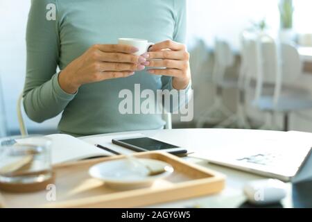 Hands of young casual female holding cup of tea over table with gadgets and open notebook while having break in cafe Stock Photo