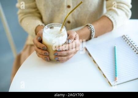 Hands of female student holding glass of cappuccino with straw while sitting by table at break and doing homework Stock Photo