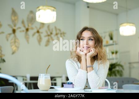 Young cheerful woman thinking of something nice while relaxing in cozy cafe by table and having cappuccino and dessert Stock Photo