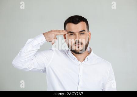 bearded man in white shirt shooting in temple with hand on gray background. Stock Photo