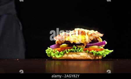 Craft burger is cooking on black background. Consist: sauce, lettuce, tomato, red onion, pickle, cheese, bacon, air bun and marbled meat beef. Stock Photo