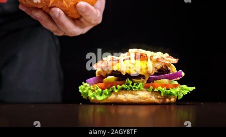 Craft burger is cooking on black background. Consist: sauce, lettuce, tomato, red onion, pickle, cheese, bacon, air bun and marbled meat beef. Stock Photo
