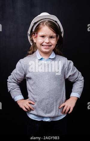 Cute little girl in hardhat and protective eyeglasses keeping hands on waist Stock Photo