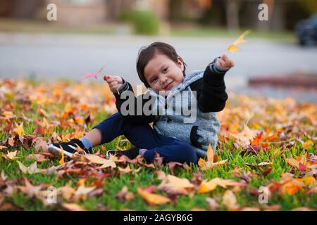 Smiling young boy wearing a letterman jacket that is sitting in the front yard, filled with yellow and red leaves from fall. Stock Photo