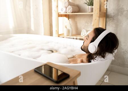 Young man with headphones lying in white bathtub filled with hot water and foam Stock Photo