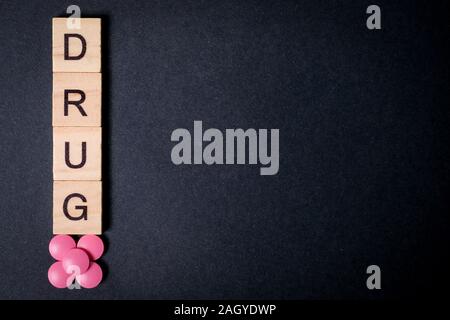 The word drug is on wooden squares next to pink tablets on a black background. Copy space. Top view. Close up. Stock Photo