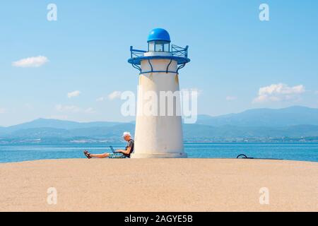 Freelancer with notebook near lighthouse in Greece. Man working on laptop near the sea. Consumers behavior. Online shopper purchases before holidays. Cost save background Stock Photo