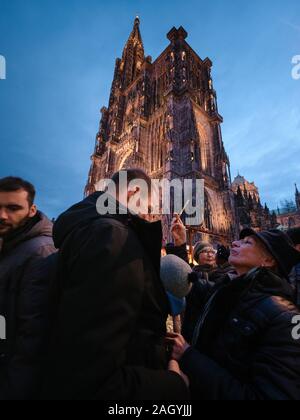 Strasbourg, France - Dec 24, 2018: Wide anlge view of happy family father child and grandmother smiling taking photos with smartphone Notre-Dame de Strasbourg cathedral in the background at dusk Stock Photo