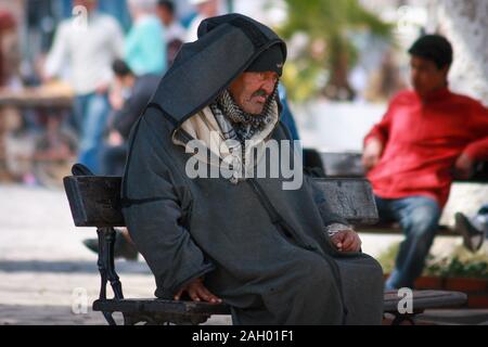 An old Tunisian man dressed in a traditional robe sitting on a bench ni the square in Sousse, Tunisia Stock Photo