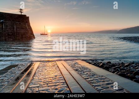 Clovelly harbour at early morning with the sun rising on the horizon and a fishing boat setting sail in this fishing village, North Devon, South West, Stock Photo