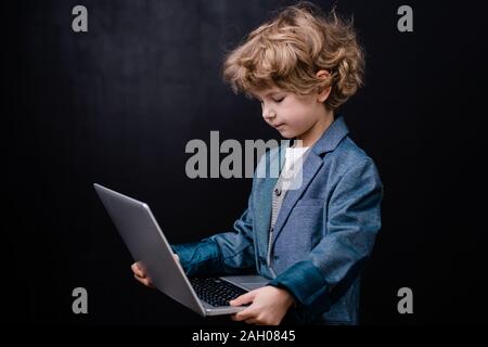 Clever elementary schoolboy holding laptop in front of himself while surfing in the net against black background Stock Photo