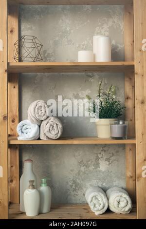 Wooden shelves with rolled towels, plastic jars with shower gel and liquid soap, green plant in flowerpot and candles against grey wall Stock Photo
