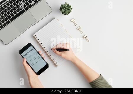 Hands of female employee holding smartphone and pen over blank page of notebook Stock Photo