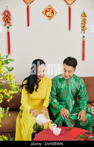 Young Vietnamese couple in traditionall dresses celebrating Lunar New Year and drinking herbal tea with dried berries Stock Photo