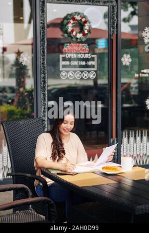 Smiling pretty young woman checking documents and taking notes in planner when having coffee at coffeeshop Stock Photo