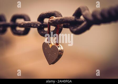 A romantic closed and rusty heart shaped padlock hanging from a chain beside a river with blurred cityscape in the background. Concept of long lasting Stock Photo