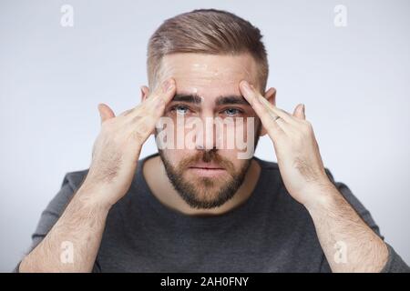 Horizontal head and shoulders portrait of young man applying nourishing face cream on his face looking at camera, white background Stock Photo
