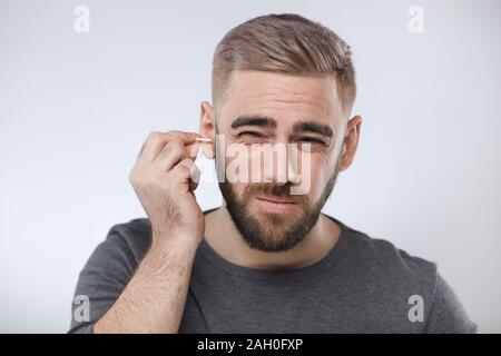 Horizontal head and shoulders portrait shot of Caucasian man wearing dark gray T-shirt cleaning his ear using cotton swab, white background Stock Photo