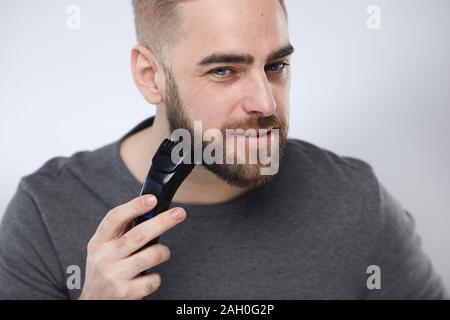Head and shoulders studio portrait shot of stylish young man grooming his beard using trimmer looking at camera Stock Photo