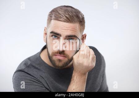 Head and shoulders studio shot of Caucasian young man tweezing his eyebrows looking at camera Stock Photo