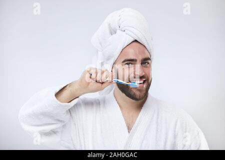 Horizontal studio portrait of cheerful young man wearing white towel headwrap and bathrobe brushing his teeth looking at camera Stock Photo