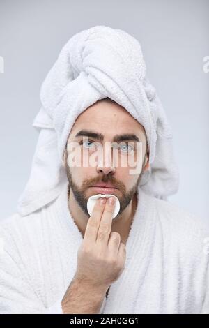 Vertical close up portrait of handsome young man applying toner on face with cotton pad looking at camera Stock Photo