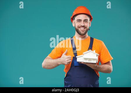 Horizontal studio portrait of construction worker posing on camera with dummy building standing against turquoise background Stock Photo