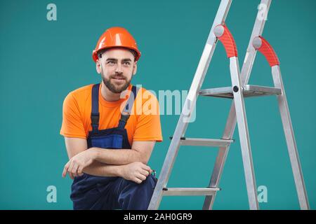 Horizontal studio portrait shot of handsome construction worker with one leg resting on ladder looking away Stock Photo
