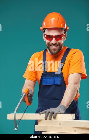 Vertical studio portrait of cheerful carpenter sawing wood planks looking at camera, blue turquoise background Stock Photo