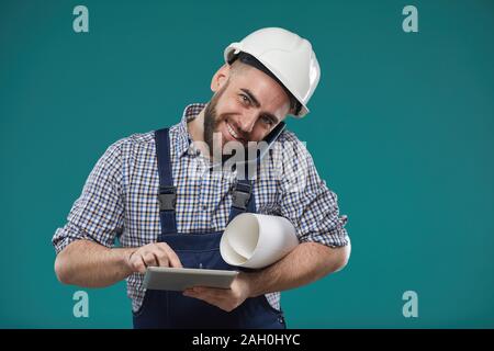 Horizontal studio shot of busy foreman holding building plan, using tablet computer and talking on phone at same time Stock Photo