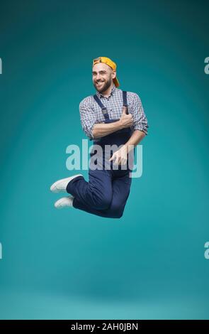 Vertical studio shot of young adult handsome man in workwear having fun jumping high with thumb up smiling Stock Photo