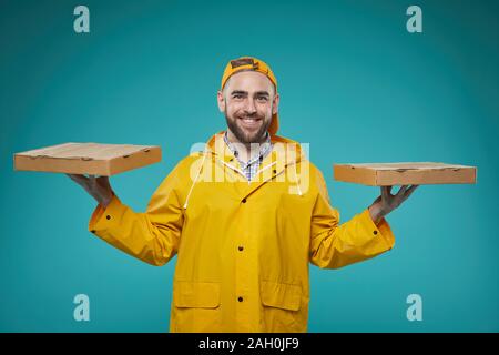 Horizontal waist up portrait of pizza man wearing bright yellow uniform holding two pizza boxes against blue turquoise wall background Stock Photo