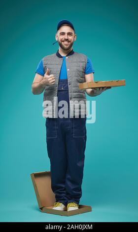 Vertical full shot studio portrait of funny delivery man standing in pizza box showing thumb up smiling at camera Stock Photo