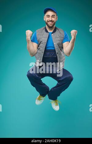 Vertical full shot of cheerful young adult man in comfortable workwear jumping against blue turquoise background Stock Photo