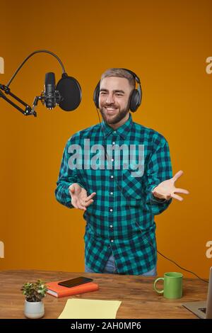 Vertical medium long shot portrait of young adult radio broadcaster wearing headphones recording content in his studio standing in front of microphone Stock Photo